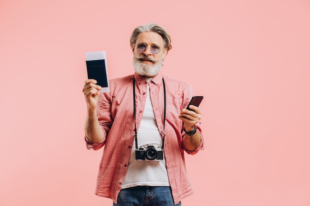 Gelukkig bebaarde man in zonnebril met een mobiele telefoon en een paspoort met kaartjes op roze