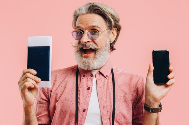 Gelukkig bebaarde man in zonnebril met een mobiele telefoon en een paspoort met kaartjes op roze