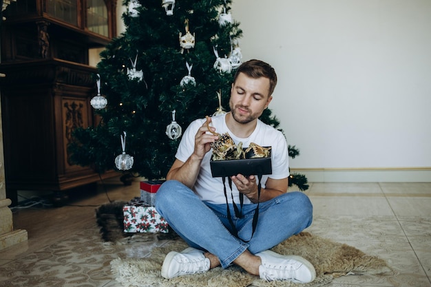 Gelukkig bebaarde man opent kerstcadeau Verrast hipster man zit in de buurt van kerstboom Man geniet thuis van kerstvakantie Gelukkig Nieuwjaar kerstcadeaus