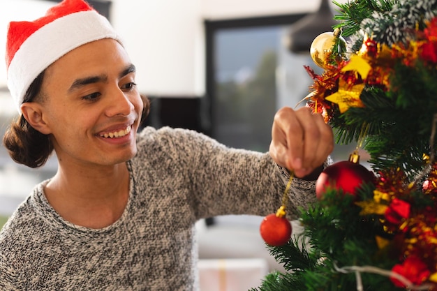 Foto gelukkig biracial man met kerstman hoed, kerstboom versieren in de woonkamer