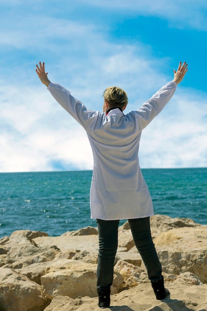 Foto gelukkig blanke amerikaanse medische vrouw in lab coat genieten van vrijheid met open handen op zee. zelfverzekerde vrouwelijke arts die zich voordeed op het strand. portret van een dokter die naar de zee loopt en zijn handen opsteekt