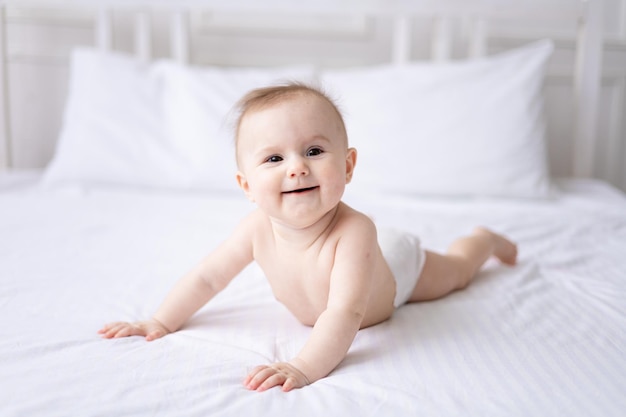 Gelukkig blanke babymeisje in wit slipje ligt op haar buik op het bed thuis in de slaapkamer op wit beddengoed kijkt naar de camera lacht gezonde baby