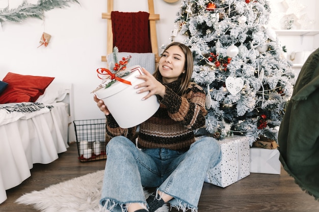 Gelukkig blanke vrouw met grote kerstcadeau zittend op de vloer thuis in de buurt van de kerstboom.