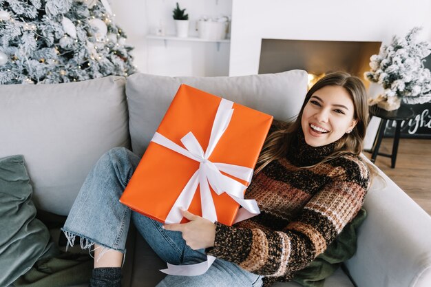 Gelukkig blanke vrouw met grote kerstcadeau zittend op een bank thuis in de buurt van de kerstboom.