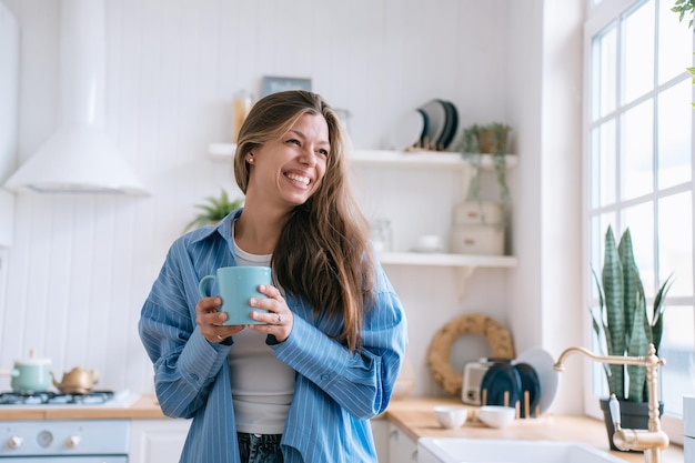 Gelukkig blanke vrouw met lang los haar in spijkerbroek en blauw shirt met een brede glimlach houdt een kopje thee vast