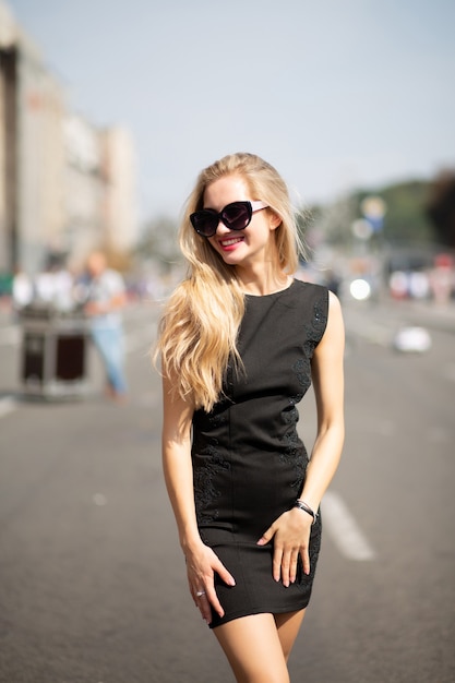 Gelukkig blond model met zwarte jurk en zonnebril, lopend door de zonnige straat