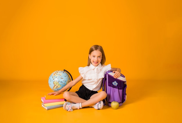 Gelukkig blond schoolmeisje in uniform zit met schoolbenodigdheden op een gele achtergrond met een kopie van de ruimte