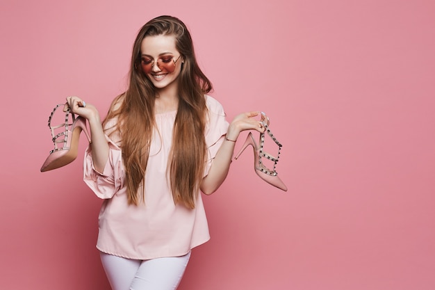 Gelukkig blonde modelmeisje met een glanzende glimlach in beige blouse en in modieuze roze zonnebril die modieuze schoenen in haar handen houden en bij de roze geïsoleerde achtergrond stellen,