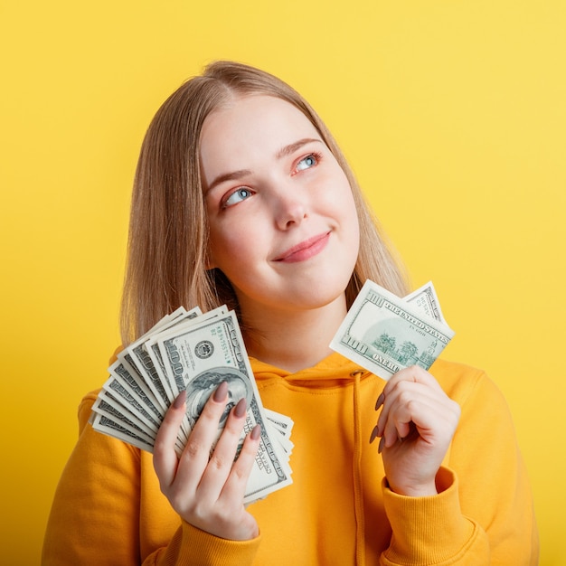 Gelukkig blonde tienermeisje in handen geld contant geld dollars dromen zorgvuldig geïsoleerd op een gele achtergrond kleur. portret jonge opgewonden lachende vrouw stapel geld bankbiljetten. close-up vierkant.