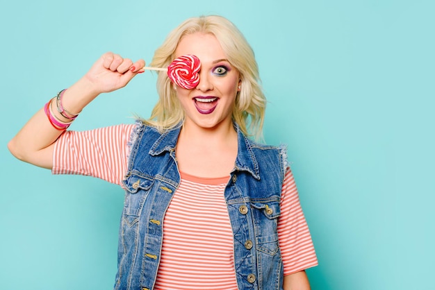 Gelukkig blonde vrouw met lolly poseren op gekleurde achtergrond. Bedek haar gezicht met lollypop