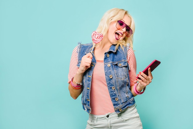 Gelukkig blonde vrouw met lolly poseren op gekleurde achtergrond. Bedek haar gezicht met lollypop