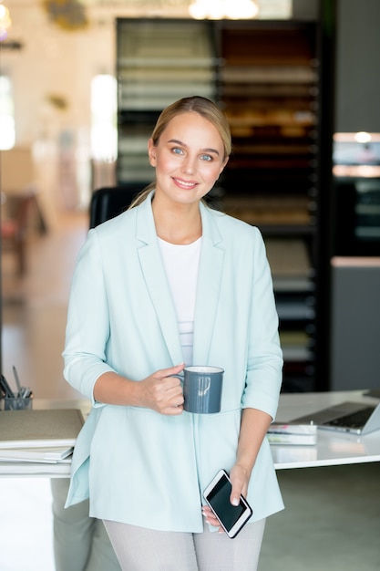 Gelukkig blonde zakenvrouw in slimme vrijetijdskleding met thee bij pauze door haar werkplek