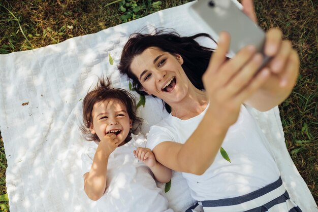 Gelukkig brunette jonge lachende vrouw zelfportret nemen naar mooi meisje dochter liggend op witte plaid op gras jong gezin selfie buiten moederschap ouderschap en jeugd concept