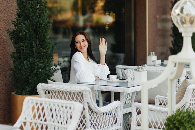 Gelukkig brunette meisje zit in een café en zwaait met haar hand naar een vriend op straat.