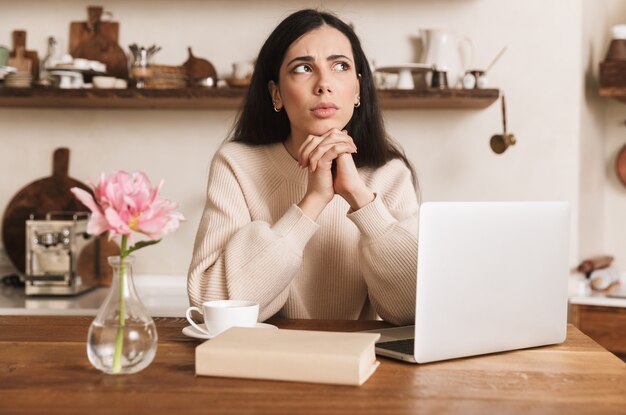 gelukkig brunette vrouw koffie drinken en laptopcomputer gebruikt in de keuken thuis