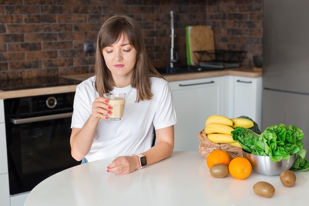 Gelukkig brunette vrouw zitten met zelfgemaakte glas smoothie en gezond fruit thuis keuken Veganistische maaltijd en detox concept Meisje met witte tshirt drinken van verse cocktail