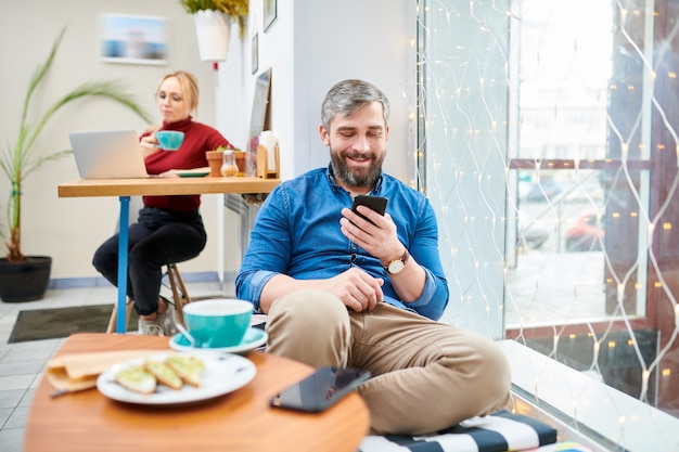 Gelukkig casual jongeman ontspannen in café terwijl scrollen in smartphone of messaging naar iemand