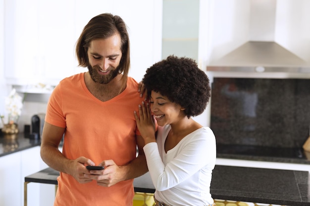 Gelukkig divers koppel in de keuken met behulp van smartphone en omarmen