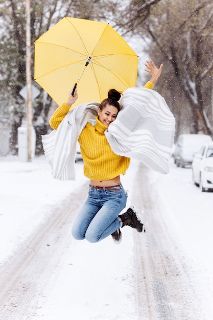 Gelukkig donkerharig meisje gekleed in een gele trui, jeans en een witte sjaal springt met een gele paraplu in een besneeuwde straat op een winterdag.