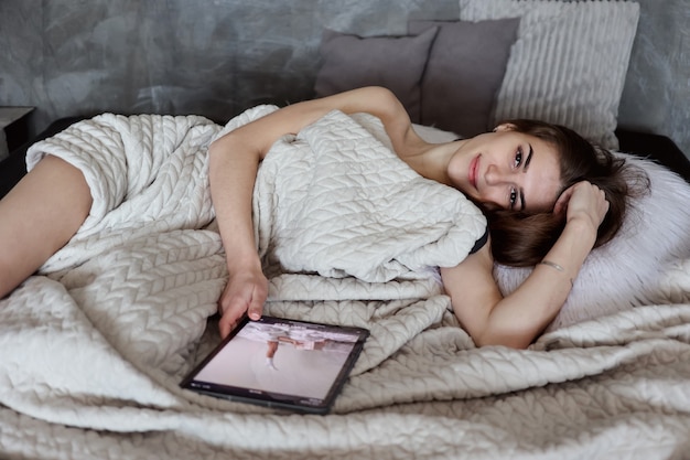 Gelukkig duizendjarige vrouw blog lezen op digitale tablet liggend in bed thuis slaapkamer