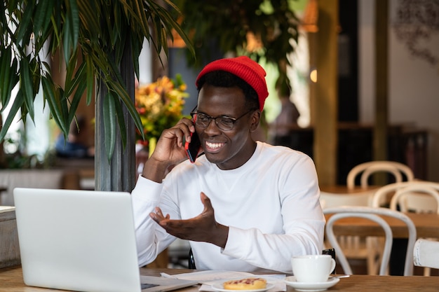 Gelukkig duizendjarige zwarte man lachen, spreken met vriend op mobiele telefoon, extern werk in café.