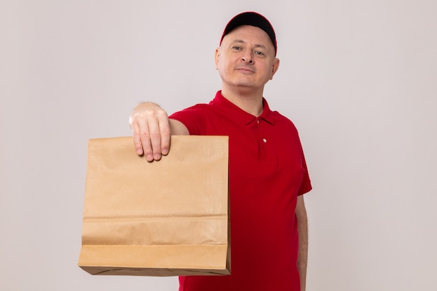 Gelukkig en positieve levering man in rood uniform en pet houden papier pakket kijken camera glimlachend zelfverzekerd staande op witte achtergrond