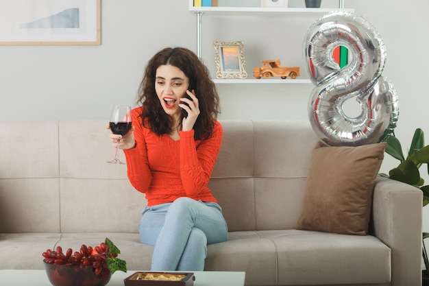Gelukkig en tevreden jonge vrouw in casual kleding glimlachend vrolijk zittend op een bank met glas wijn praten op mobiele telefoon in lichte woonkamer vieren internationale vrouwendag 8 maart