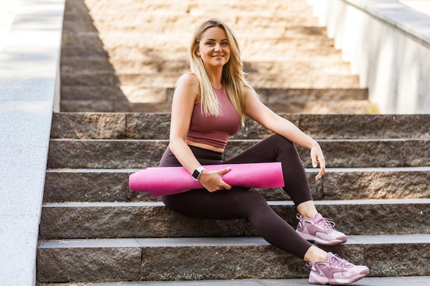 Gelukkig fit blond meisje in yogabroek zittend op trappen, mat vasthoudend en glimlachend met tevreden tevreden uitdrukking, ontspannend pauze nemen na sporttraining buiten. Genieten van een gezonde actieve levensstijl