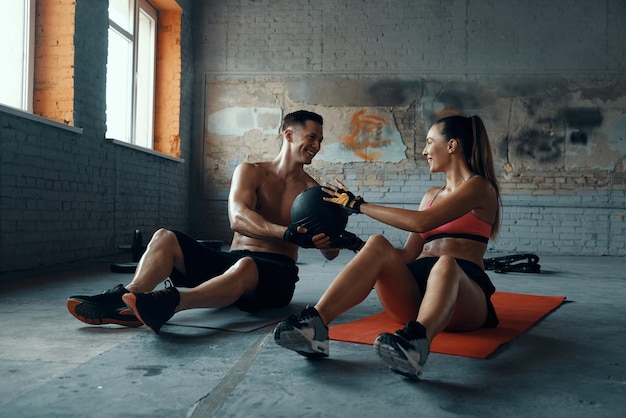 Foto gelukkig fit koppel oefent met een medicijnbal terwijl ze op oefenmatten in de sportschool zitten