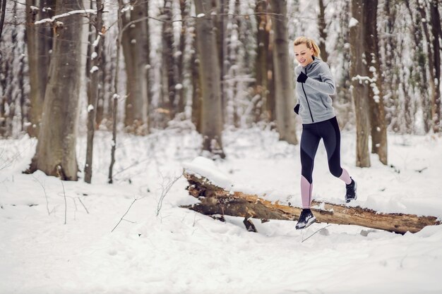 Gelukkig fit vrouw joggen in het bos op besneeuwde winterdag en springen over het gevallen hout. Gezonde levensstijl, obstakel, succes