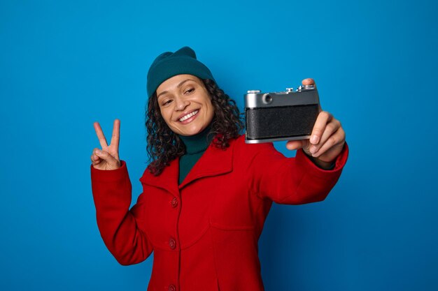 Gelukkig fotograaf, aantrekkelijke lachende Afrikaanse vrouw in rode jas en groene hoed vredesteken tonen terwijl het nemen van een zelfportret met een vintage camera, geïsoleerd op blauwe achtergrond met kopie advertentieruimte