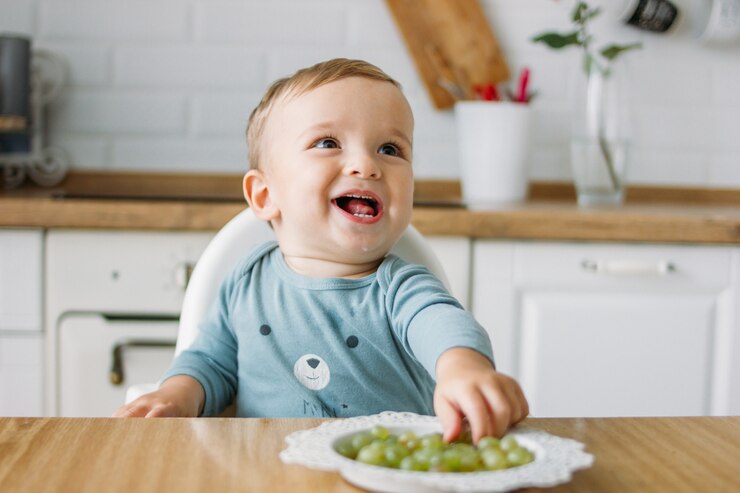 Gelukkig gelukkig weinig babyjongen die eerste voedsel groene druif thuis eten bij de heldere keuken