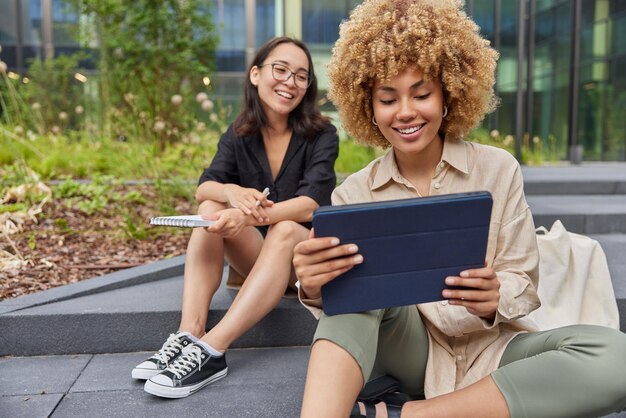 Gelukkig gemengd ras vrouwelijke vrienden ontmoeten elkaar nemen selfie op moderne tablet voorbereiden op examens gekleed in vrijetijdskleding zitten op trappen buiten in de stad bladeren touchpad kijken naar online filminhoud