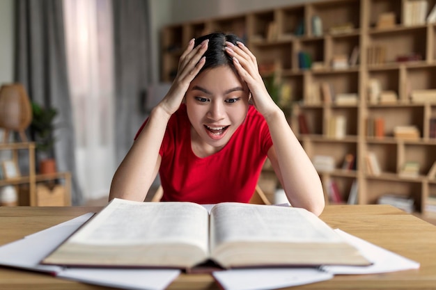 Gelukkig geschokte slimme jonge Koreaanse vrouwelijke student die naar een boek kijkt, geniet van onderwijs in het interieur van de kamer