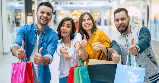 Gelukkig gezelschap van lachende opgewonden mooie stijlvolle vrienden met kleurrijke papieren zakken hebben plezier en duimen opdagen tijdens het winkelen in het winkelcentrum.