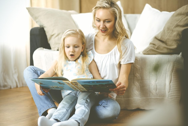 Gelukkig gezin. Blonde jonge moeder die een boek leest aan haar schattige dochter terwijl ze op een houten vloer zit. Moederschap concept