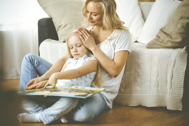 Gelukkig gezin. Blonde jonge moeder leest een boek voor aan haar schattige dochter terwijl ze op de houten vloer zit. Moederschap concept.