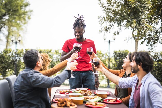 Gelukkig gezin juichen met rode wijn bij barbecue lunch buiten verschillende leeftijden van mensen die plezier hebben in het weekend maaltijd eten smaak en familie concept focus op afrikaanse jonge man