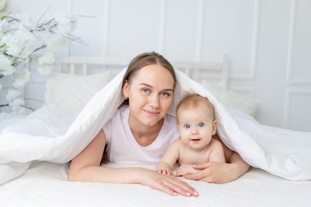 Gelukkig gezin, moeder met een lachende baby onder een deken op een wit katoenen bed thuis knuffelen