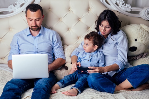 Gelukkig gezin, mooie moeder zoontje rust online in de telefoon en vader zakenman werkt samen in een laptop op bed in de slaapkamer