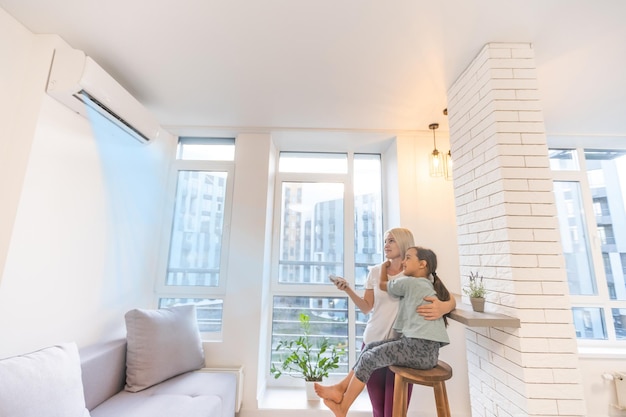 Foto gelukkig gezin onder airconditioner, moeder met afstandsbedieningschakelaar op airconditioning in woonkamer pas comforttemperatuur aan voor dochter, klimaatsysteem in modern huis