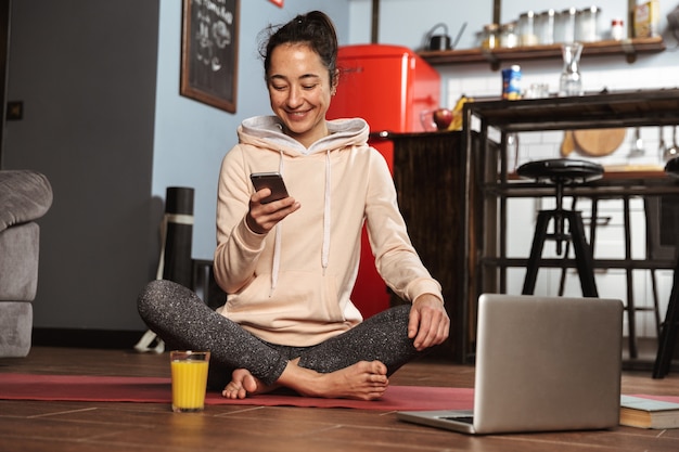 Gelukkig gezonde vrouw zittend op een fitness-mat en thuis praten over de mobiele telefoon