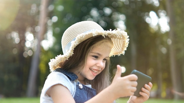 Gelukkig glimlachend kindmeisje dat in de zomer buiten op haar mobiele telefoon kijkt