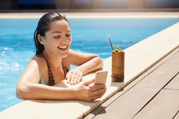 Gelukkig glimlachend wijfje die met nat haar moderne slimme telefoon in hand houden, bij poolside ontspannen en niet-alcoholische cocktail drinken