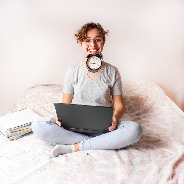 Gelukkig grappig tienermeisje die op het bed op computer bestuderen en wekker op haar mond houden