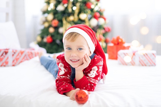 Gelukkig grappige babyjongen met geschenken in bed op kerstochtend op de achtergrond van de kerstboom, het concept van vakantie, kerstmis, nieuwjaar