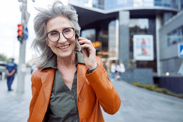 Gelukkig grijsharige senior vrouw met bril praat op mobiele telefoon op straat in de stad