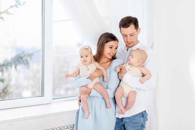 Gelukkig groot gezin met een tweeling, mama en papa met twee pasgeboren baby-tweelingmeisjes in een katoenen pak bij het raam van het huis, ze knuffelen en kussen, moederschap en vaderschap met twee kinderen