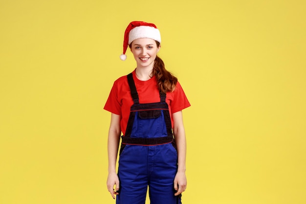 Gelukkig handige vrouw in blauwe overalls en kerstmuts staande kijken camera wachtvolgorde beroep van service industrie huis reparatie Indoor studio opname geïsoleerd op gele achtergrond