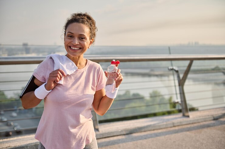 Foto gelukkig heerlijke afro-amerikaanse sportieve vrouw jogger met een fles met water en een witte handdoek op haar schouders, glimlacht met mooie brede glimlach kijken naar camera staande op een loopband